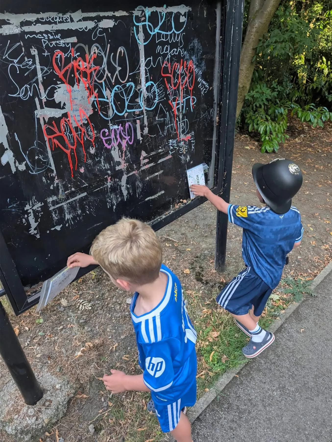 Two young boys in blue soccer jerseys and shorts are cleaning graffiti off a black signboard with white sponges.