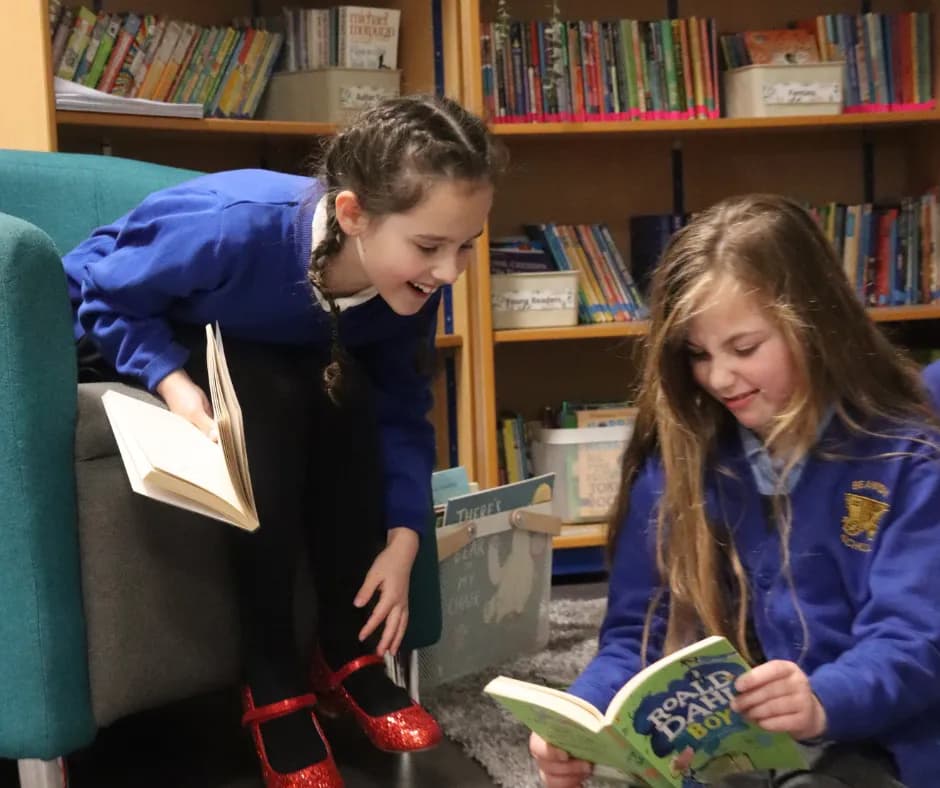 Two young girls in blue sweaters are reading books, one sitting on a chair and the other on the floor.